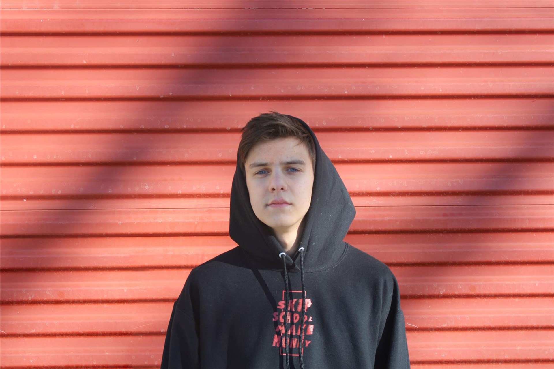 Ein Junge von ca. 13 Jahren mit schwarzen Pullover und Kapuze steht vor einer roten Wand und schaut in die Kamera, der Blick ernst mit nur leichtem Lächeln