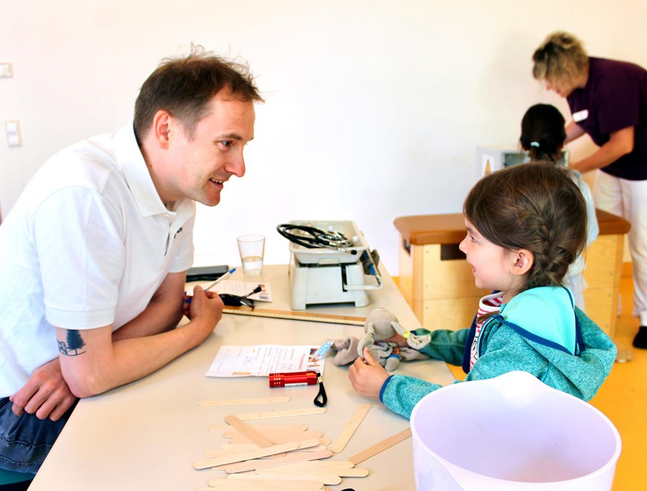Freundlich zugewandte Person an einem Schreibtisch im Gespräch mit gegenüber sitzendem Kind mit Kuscheltier.