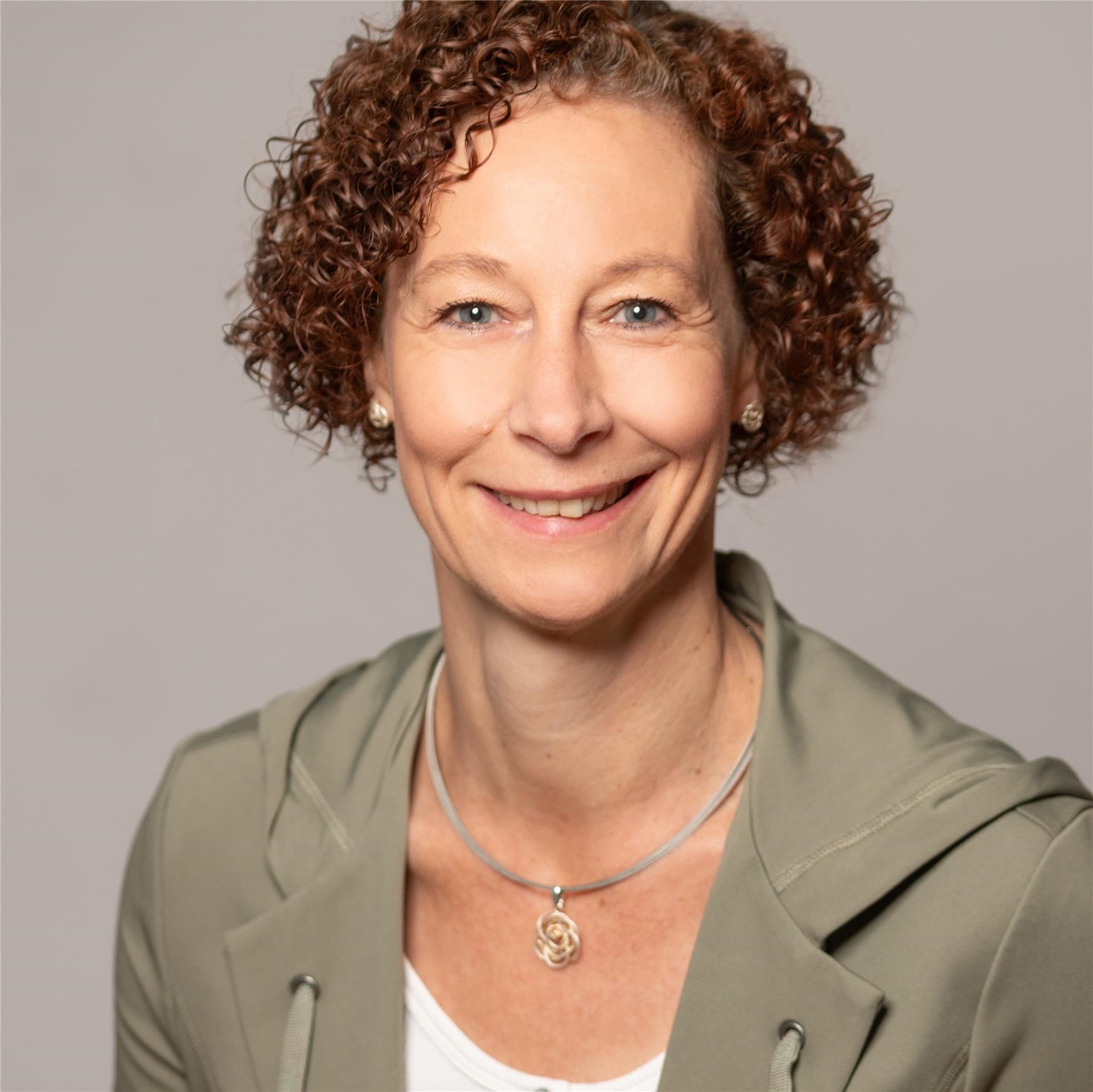 Frau mit kurzen, lockigen roten Haaren trägt olivgrüne Jacke, weiße Bluse und silberne Kette mit rundem Anhänger.
